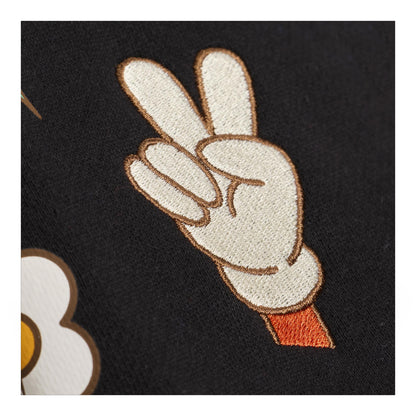 Close-up of a black garment featuring an embroidered cartoon hand making a peace sign in off-white with brown outlines and a small orange cuff, alongside part of a white flower motif.